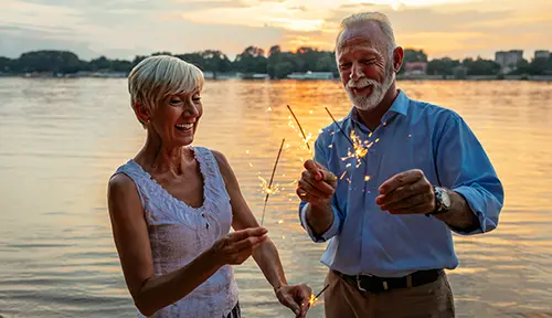 Two people holding sparklers by a body of water at sunset.